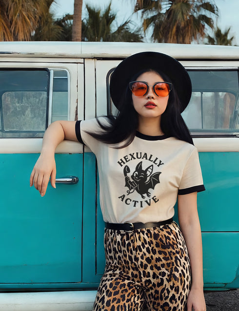 Unisex offwhite and black ringer t-shirt with a bold black graphic of a cartoon bat holding a wand, surrounded by the phrase “Hexually Active.” The design is playful, witchy, and gothic, perfect for lovers of magic, spellwork, hexes, and witch humor. The shirt features contrast collar and sleeves in black for a retro vibe.