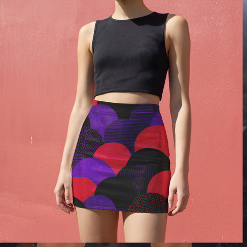 A woman wearing a black crop top and a colorful skirt with a pattern of circles and lines on it, standing against a pink wall.