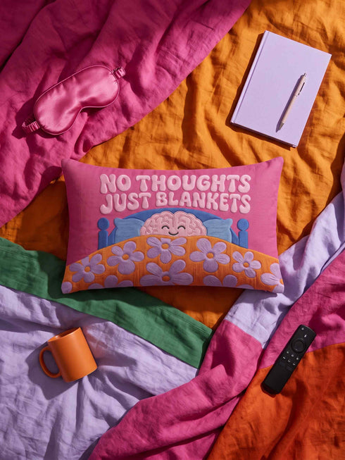 A bright magenta pink rectangular lumbar pillow cover featuring a flat 2D printed faux embroidery design. The printed illusion shows light pink bubble text reading NO THOUGHTS JUST BLANKETS. Below the text is a cartoon pink brain with a happy face, tucked into a blue bed with blue pillows. The brain is covered by a textured-looking orange blanket decorated with light purple daisies. The entire design is a smooth printed trompe l'oeil stitch effect, not real woven or stitched texture.