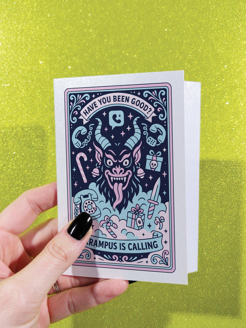 A vertical 5x7 holiday card featuring a retro tattoo style illustration of Krampus in pastel pink, teal, and black ink on shimmery silver pearlescent paper. The central horned demon has a long tongue and is surrounded by vintage rotary phones, daggers, candy canes, and skull gifts. Top text asks Have You Been Good while the bottom banner reads Krampus Is Calling. The interior text reads Let's be honest... Being good is overrated. Merry Christmas.