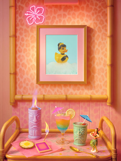 A chic rubber duck styled like Audrey Hepburn’s Holly Golightly, wearing a high bun, yellow bow, black gloves, and a sparkly choker. The duck floats in a pile of foam and is framed in white on a bubbly bathroom shelf with a red tile backdrop and soap dispenser.