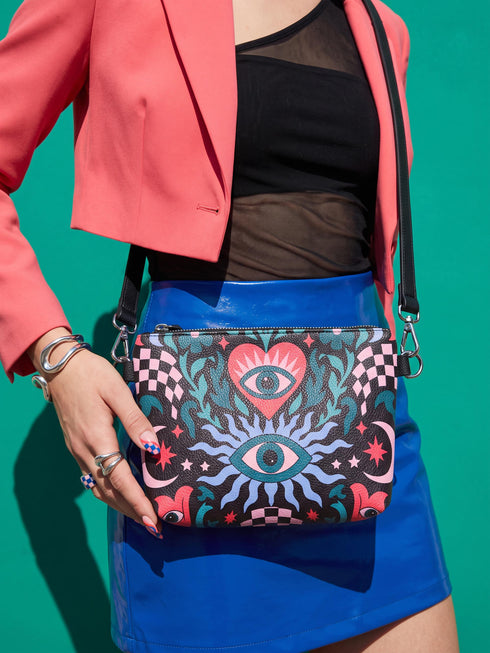 Small vegan leather crossbody purse with a real pebble grain texture and adjustable black strap. The flat printed artwork features a central blue sunburst evil eye motif below a coral pink sacred heart eye, surrounded by pink crescent moons, multicolored stars, flowing teal botanical vines, and wavy lavender and black checkerboard distortion banners. Set against a dark background with silver metal hardware including a top zipper, zipper pull, and strap clips. Measures 8.5 by 7 by 2 inches.
