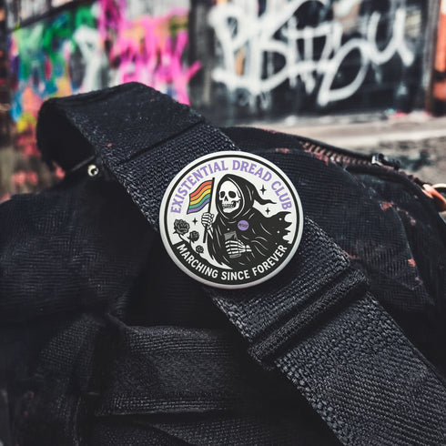 Round pinback button featuring a bold black and white design of a skeleton reaper holding a rainbow pride flag and a drink, wearing a "RESIST" badge, surrounded by black roses, with "Existential Dread Club" in purple letters and "Marching Since Forever" in white. Glossy, metal button in 2" or 3" size.