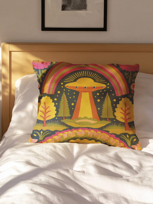 A colorful, patterned pillow with a whimsical design sits on a bed, featuring a rainbow, trees, and an alien-like figure.