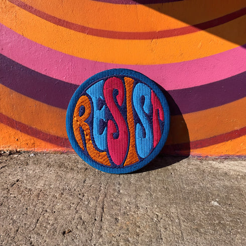 Round embroidered iron-on patch featuring "RESIST" text in bold flame design. Blue border surrounds navy background with orange, red, and blue flame-style lettering. 3x3 inch political activist patch with premium twill fabric construction, perfect for jackets, backpacks, and clothing customization. Anti-establishment protest accessory for resistance movements and political activism demonstrations.