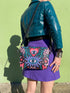Small vegan leather crossbody purse with a real pebble grain texture and adjustable black strap. The flat printed artwork features a central blue sunburst evil eye motif below a coral pink sacred heart eye, surrounded by pink crescent moons, multicolored stars, flowing teal botanical vines, and wavy lavender and black checkerboard distortion banners. Set against a dark background with silver metal hardware including a top zipper, zipper pull, and strap clips. Measures 8.5 by 7 by 2 inches.