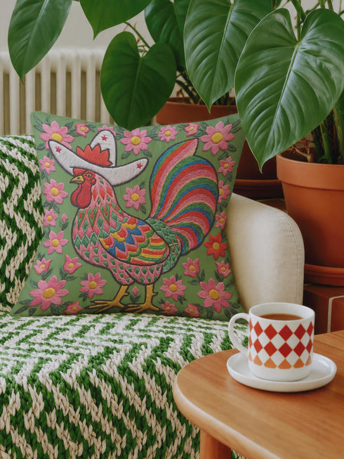 Square throw pillow cover with a printed faux embroidery design of a rooster wearing a white cowboy hat with red stars on a grass green background. The western chicken has a red comb and wattle, yellow beak and feet, and layered feathers in pink, green, orange, blue, and yellow with a rainbow striped tail. Pink and red flowers with yellow centers frame the bird. The stitched look is a flat 2D print on a smooth linen feel polyester cover with a hidden zipper, not raised embroidery.