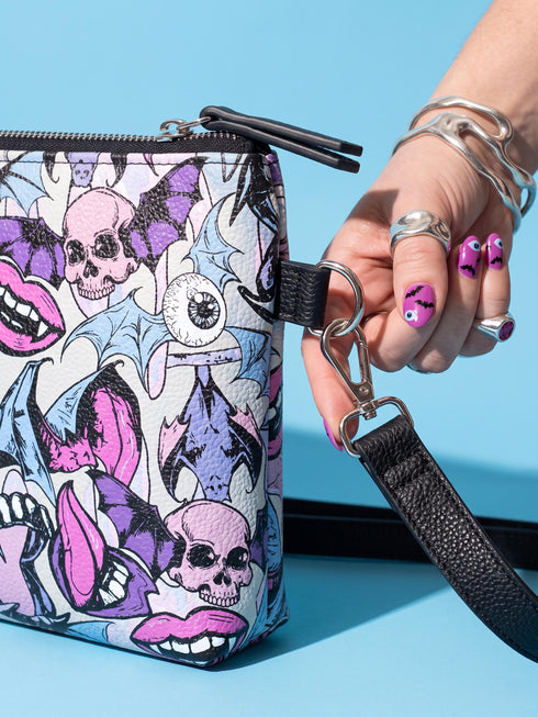Small vegan leather crossbody purse with a real pebble grain texture and adjustable black strap. The flat printed artwork features a scattered all over pattern of pink skulls, purple and pastel blue winged bats, pink fanged mouths, and loose eyeballs set against a cream colored background. The bag is shown with black and silver hardware including a black top zipper, black zipper pull, black strap tabs, and silver metal rings and clips. The compact rectangular bag measures 8.5 by 7 by 2 inches.