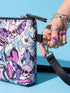 Small vegan leather crossbody purse with a real pebble grain texture and adjustable black strap. The flat printed artwork features a scattered all over pattern of pink skulls, purple and pastel blue winged bats, pink fanged mouths, and loose eyeballs set against a cream colored background. The bag is shown with black and silver hardware including a black top zipper, black zipper pull, black strap tabs, and silver metal rings and clips. The compact rectangular bag measures 8.5 by 7 by 2 inches.