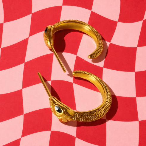 a pair of gold snake-shaped earrings with black stones on a red and white checkered background.