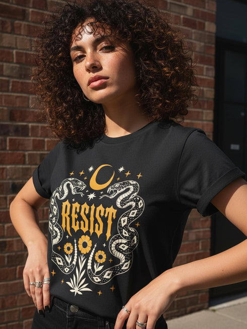 A unisex short sleeve graphic t shirt in dark charcoal black featuring a highly detailed symmetrical medallion print on the chest. In the center, the word RESIST is printed in a bold yellow gothic blackletter font. Two coiled white snakes with fine black scale details frame the text on the left and right. Above the text, a yellow crescent moon is flanked by white and yellow starbursts. Beneath, three yellow marigold flowers sit atop white stems and leafy foliage.