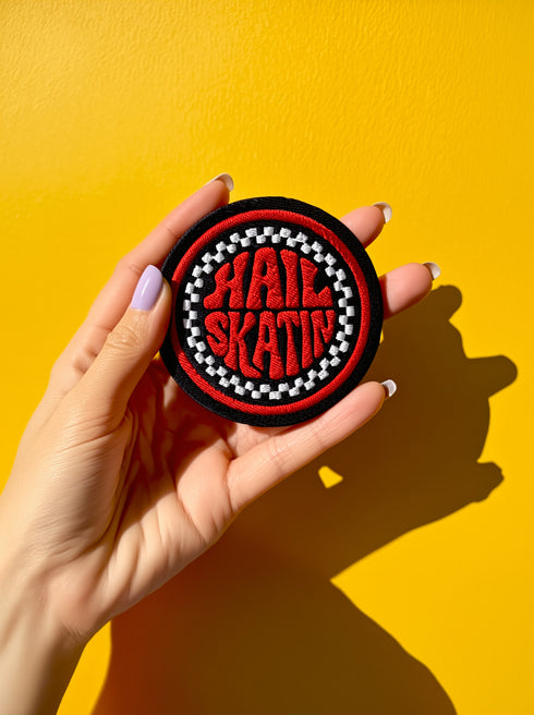 A round, black embroidered patch with a thick, textured merrow border stitched in alternating orange and yellow thread. In the center, the words "PAPERBACK WITCH" are embroidered in a retro 70s-style font. The word "PAPERBACK" is arched across the top in bold orange thread with a thin yellow outline. The word "WITCH" is across the bottom in puffy, bright white thread with a thin yellow outline. Between the words is a central icon of an open book, containing a stylized all-seeing eye.