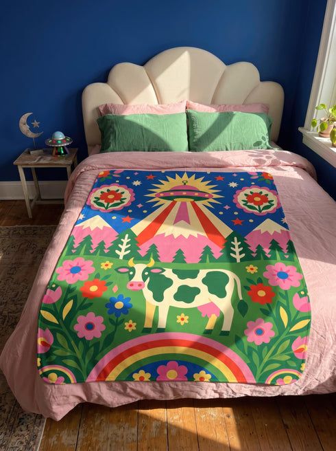 A rectangular velveteen minky blanket with a bright, maximalist design. The top half features a vibrant blue sky with a pink, green, and yellow UFO beaming rays down over hot pink snow-capped mountains. The bottom half shows a grassy green field with a white cow covered in dark spots, surrounded by large, colorful red, pink, and blue retro flowers, pine trees, and a rainbow arch at the very bottom. The reverse side is a fluffy off-white sherpa.