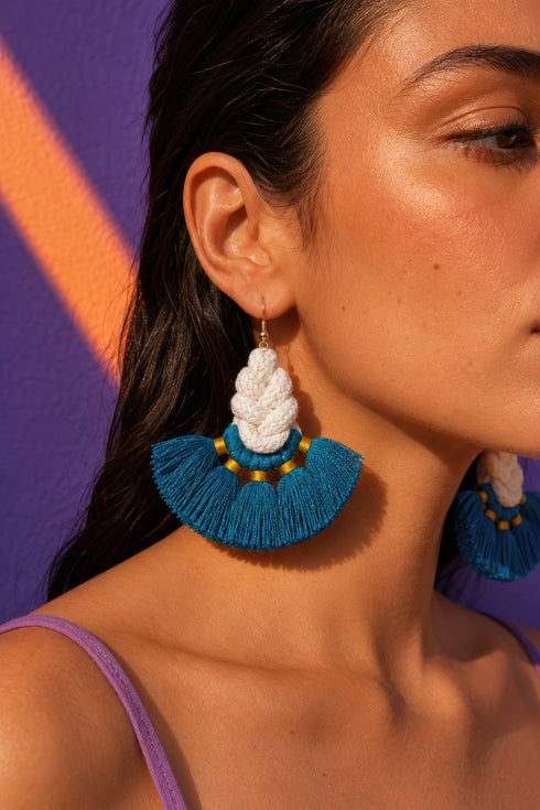 A woman wearing large, blue and white earrings with tassels, set against a purple background.