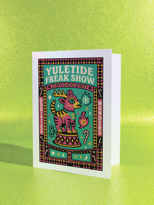Maximalist Yuletide Freak Show 5x7 folded greeting card with a pearlescent shimmer finish. Features a vibrant, retro circus illustration in teal, pink, and yellow. The central motif is a bizarre, striped reindeer-tiger hybrid with a wind-up key, standing on a pedestal. The graphic border contains 'YULETIDE FREAK SHOW,' 'ODDITIES,' and 'WONDERS.' This unique and witty card is an unconventional alternative Christmas gift for oddity collectors.