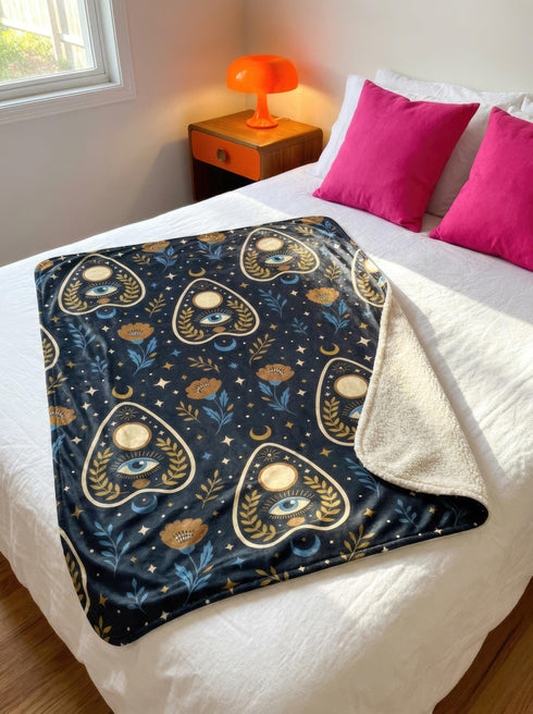 A rectangular velveteen minky blanket featuring a repeating dark academia pattern on a deep black background. The design includes gold and cream Ouija planchettes, each containing a blue all-seeing eye, a cream circle, and a light blue crescent moon framed by botanical leaves. Scattered between the planchettes are gold flowers with light blue stems, additional crescent moons, and small four-point stars. The reverse side of the blanket is made of a fluffy, textured off-white sherpa fleece.