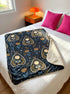A rectangular velveteen minky blanket featuring a repeating dark academia pattern on a deep black background. The design includes gold and cream Ouija planchettes, each containing a blue all-seeing eye, a cream circle, and a light blue crescent moon framed by botanical leaves. Scattered between the planchettes are gold flowers with light blue stems, additional crescent moons, and small four-point stars. The reverse side of the blanket is made of a fluffy, textured off-white sherpa fleece.