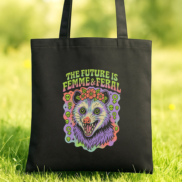 A black tote bag with a colorful graphic design featuring a cartoon-like creature with a flower crown and the text "THE FUTURE IS FEMME & FERAL" printed on it.