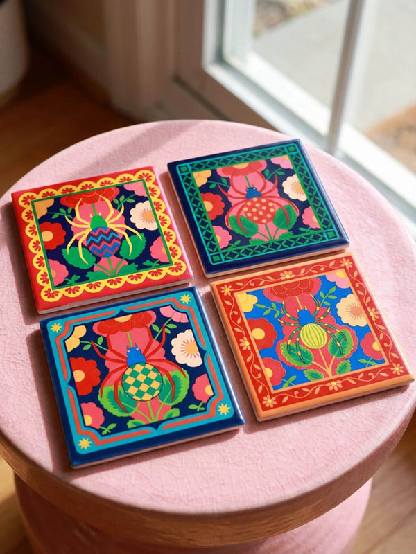 A set of four square coasters featuring a glossy glazed finish and vibrant insect illustrations. Each coaster measures approximately four by four inches. The designs showcase symmetrical beetles and spiders in bold red, blue, and orange colors, framed by intricate floral and geometric borders in a maximalist folk art style. Available in ceramic with cork backing or hardboard MDF. The edges are smooth. The designs are vivid and highly saturated, designed to provide grip and protect furniture.