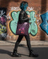 A person with blue hair is walking down a street, carrying a purple tote bag with a skateboard design on it. The background features a brick wall with graffiti.