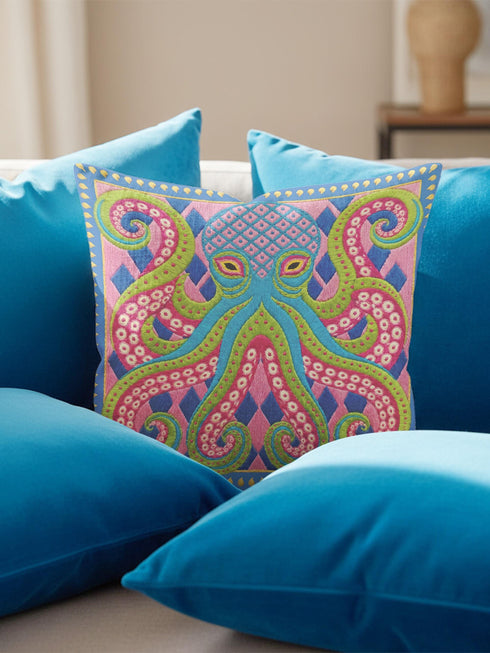 A square decorative throw pillow cover featuring a highly symmetrical bright octopus design. The octopus has a turquoise blue body with a diamond patterned head and eight curled tentacles detailed in bright pink and lime green. The background is a repeating diamond argyle pattern in royal blue and light pink. The artwork is a flat two dimensional print that mimics the textured look of intricate crewelwork embroidery. The pillow features a hidden zipper on the bottom edge. No insert included.