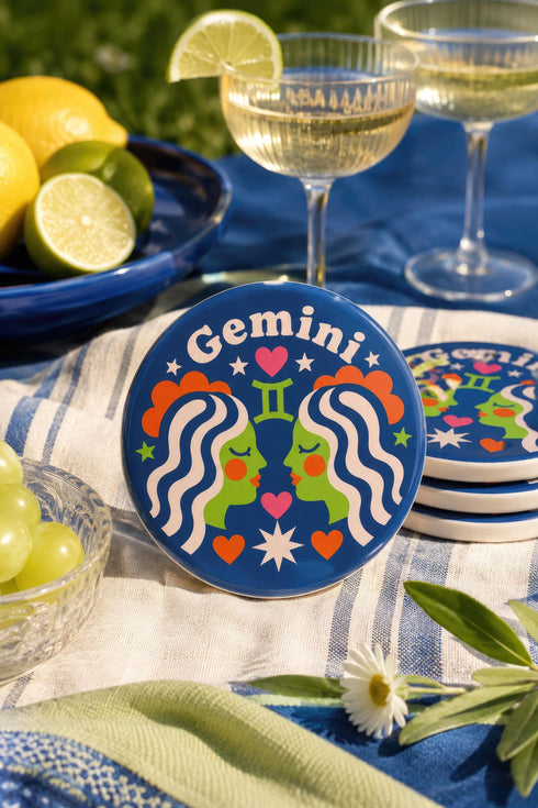 A close-up photograph of a round, glossy ceramic coaster featuring a bold retro pop art design. The background is a deep royal blue. In the center are two symmetrical profile faces of twins looking at each other, featuring bright green skin, closed eyes, and red cheeks. They have wavy white and pale pink hair with red accents. The word Gemini is arched at the top in a retro white font. The design is decorated with a green Gemini symbol, pink and red hearts, and white starbursts.