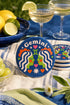 A close-up photograph of a round, glossy ceramic coaster featuring a bold retro pop art design. The background is a deep royal blue. In the center are two symmetrical profile faces of twins looking at each other, featuring bright green skin, closed eyes, and red cheeks. They have wavy white and pale pink hair with red accents. The word Gemini is arched at the top in a retro white font. The design is decorated with a green Gemini symbol, pink and red hearts, and white starbursts.