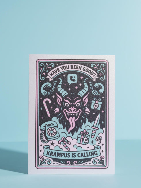 A vertical 5x7 holiday card featuring a retro tattoo style illustration of Krampus in pastel pink, teal, and black ink on shimmery silver pearlescent paper. The central horned demon has a long tongue and is surrounded by vintage rotary phones, daggers, candy canes, and skull gifts. Top text asks Have You Been Good while the bottom banner reads Krampus Is Calling. The interior text reads Let's be honest... Being good is overrated. Merry Christmas.