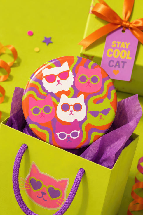 A close-up photograph of a glossy round ceramic coaster featuring a retro 70s psychedelic design. The artwork displays a cluster of six stylized cat face illustrations, each wearing a different pair of sunglasses including retro round and cat-eye shapes. The cats are colored in solid shades of white, hot pink, purple, and lime green. They are set against a vibrant background of wavy, swirling lines in bright orange, red, and purple. The top surface has a highly reflective clear glaze.