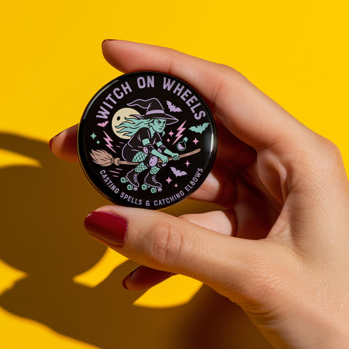 A hand holding a round pin with a witch on wheels design against a yellow background.