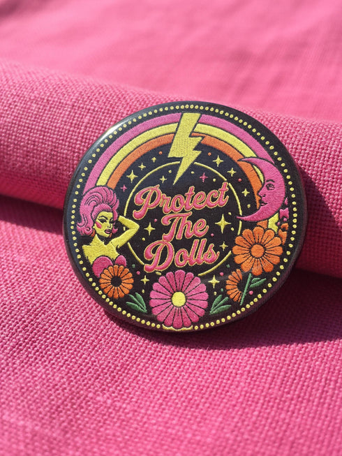 A circular black pinback button featuring the slogan Protect The Dolls in retro script. The design utilizes a high-fidelity trompe-l'œil print style that mimics the texture of raised thread embroidery with satin-stitch detailing. The artwork features vibrant hot pink, rust orange, and golden yellow motifs including drag queens with bouffant hair, 70s style daisies, lightning bolts, and a crescent moon with a face. The surface is smooth and glossy, creating a faux-textured illusion.