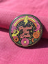 A circular black pinback button featuring the slogan Protect The Dolls in retro script. The design utilizes a high-fidelity trompe-l'œil print style that mimics the texture of raised thread embroidery with satin-stitch detailing. The artwork features vibrant hot pink, rust orange, and golden yellow motifs including drag queens with bouffant hair, 70s style daisies, lightning bolts, and a crescent moon with a face. The surface is smooth and glossy, creating a faux-textured illusion.