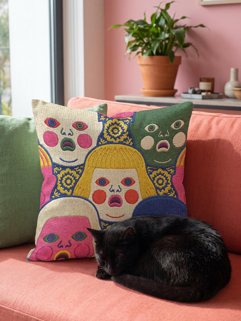 A square decorative pillow cover featuring a faux embroidery pattern of five stylized, expressive doll faces. The central face has yellow hair and pink cheeks. It is surrounded by four other faces in shades of pale beige, green, and bright pink, all with wide staring eyes and open mouths. The design uses intricate, colorful line work to create the illusion of thick woven stitching, but the fabric is a smooth print. Medallions fill the gaps.