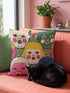 A square decorative pillow cover featuring a faux embroidery pattern of five stylized, expressive doll faces. The central face has yellow hair and pink cheeks. It is surrounded by four other faces in shades of pale beige, green, and bright pink, all with wide staring eyes and open mouths. The design uses intricate, colorful line work to create the illusion of thick woven stitching, but the fabric is a smooth print. Medallions fill the gaps.