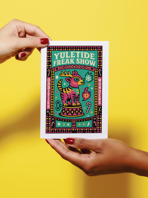 Maximalist Yuletide Freak Show 5x7 folded greeting card with a pearlescent shimmer finish. Features a vibrant, retro circus illustration in teal, pink, and yellow. The central motif is a bizarre, striped reindeer-tiger hybrid with a wind-up key, standing on a pedestal. The graphic border contains 'YULETIDE FREAK SHOW,' 'ODDITIES,' and 'WONDERS.' This unique and witty card is an unconventional alternative Christmas gift for oddity collectors.