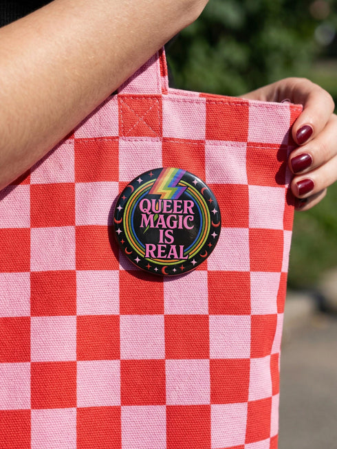 A round metal pinback button badge featuring a black background with the words QUEER MAGIC IS REAL in capital pink retro serif typography. A bright rainbow colored lightning bolt strikes from the top center, surrounded by concentric rainbow rings in red, orange, yellow, green, blue, and purple. The design is accented with small pink four-point stars and red crescent moons. The artwork features a realistic 2-D faux embroidery texture. The back has a silver metal safety pin clasp fastening.