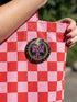 A round metal pinback button badge featuring a black background with the words QUEER MAGIC IS REAL in capital pink retro serif typography. A bright rainbow colored lightning bolt strikes from the top center, surrounded by concentric rainbow rings in red, orange, yellow, green, blue, and purple. The design is accented with small pink four-point stars and red crescent moons. The artwork features a realistic 2-D faux embroidery texture. The back has a silver metal safety pin clasp fastening.