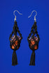 A pair of black earrings with orange pots and tassels hanging from the ends, set against a bright blue background.