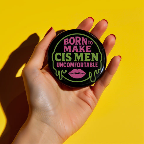 Glossy black pinback button featuring bold text 'Born to Make Cis Men Uncomfortable' in pink and green glitter fonts. Design includes melting skull outline and lips illustration. Button is round and 2.25 inches in diameter with a durable tinplate body and UV-resistant coating. Perfect for jackets, backpacks, or gifts for queer punks and feminists.