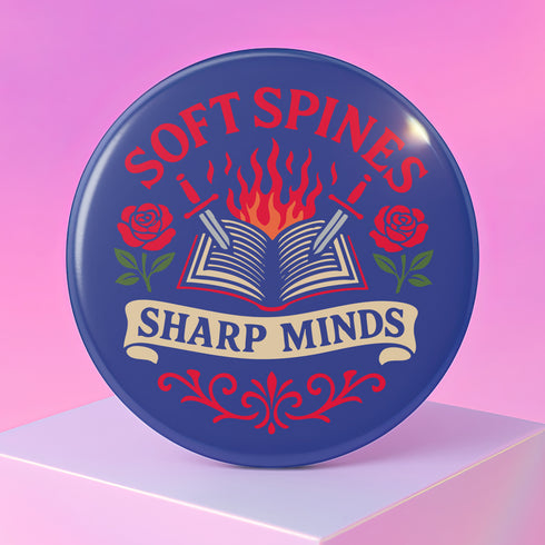 A round pinback button in blue with a glossy mylar face and metal construction. The design, in a traditional tattoo style, features a central open book with flames rising from its pages. Two silver swords with black hilts are crossed over the book. On either side of the book is a red rose with a green stem and leaves. Below the book is a cream-colored banner with the words "SHARP MINDS" in a black serif font. Above the book, the words "SOFT SPINES" are written in a curved, red font.