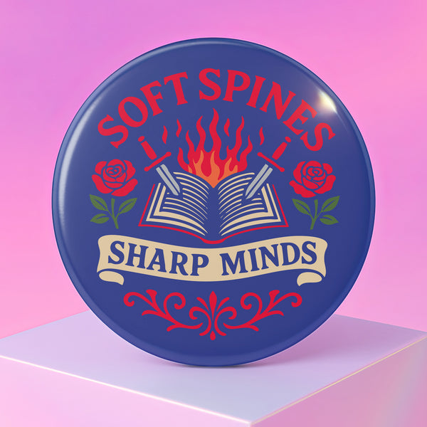 A round pinback button in blue with a glossy mylar face and metal construction. The design, in a traditional tattoo style, features a central open book with flames rising from its pages. Two silver swords with black hilts are crossed over the book. On either side of the book is a red rose with a green stem and leaves. Below the book is a cream-colored banner with the words "SHARP MINDS" in a black serif font. Above the book, the words "SOFT SPINES" are written in a curved, red font.