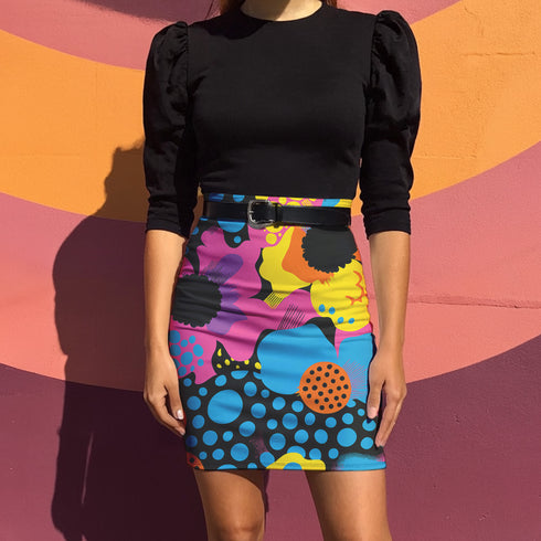 A woman wearing a black top and a colorful, patterned skirt stands in front of a vibrant, abstract wall.