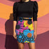 A woman wearing a black top and a colorful, patterned skirt stands in front of a vibrant, abstract wall.