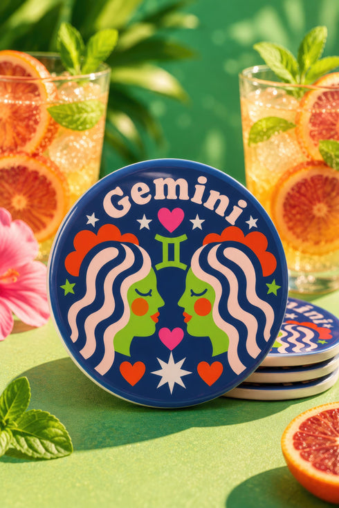 A close-up photograph of a round, glossy ceramic coaster featuring a bold retro pop art design. The background is a deep royal blue. In the center are two symmetrical profile faces of twins looking at each other, featuring bright green skin, closed eyes, and red cheeks. They have wavy white and pale pink hair with red accents. The word Gemini is arched at the top in a retro white font. The design is decorated with a green Gemini symbol, pink and red hearts, and white starbursts.
