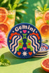 A close-up photograph of a round, glossy ceramic coaster featuring a bold retro pop art design. The background is a deep royal blue. In the center are two symmetrical profile faces of twins looking at each other, featuring bright green skin, closed eyes, and red cheeks. They have wavy white and pale pink hair with red accents. The word Gemini is arched at the top in a retro white font. The design is decorated with a green Gemini symbol, pink and red hearts, and white starbursts.