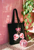 Black 15 x 16 inch 100% cotton tote bag with a kawaii pastel goth cherry-skull design printed on the front. The artwork features two soft pink skulls shaped like cherries, connected by red stems and topped with a bright pink bow. Cute, spooky, coquette, and alt-inspired everyday tote.