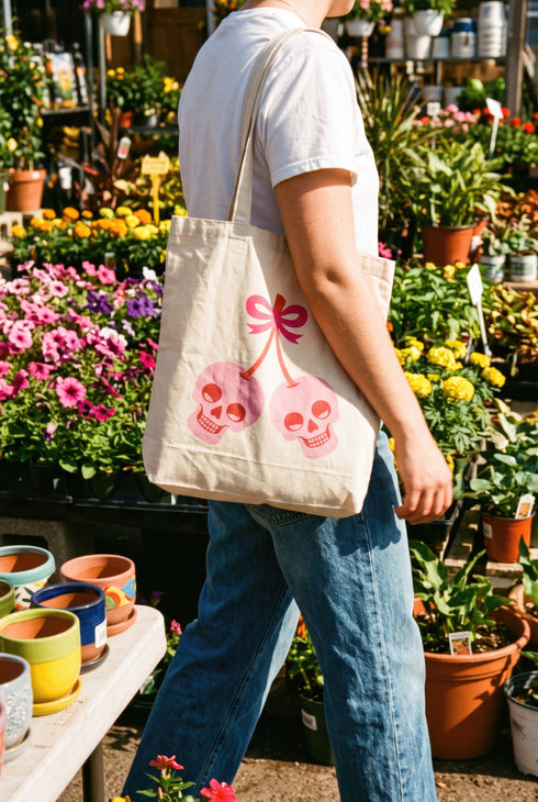 White 15 x 16 inch 100% cotton tote bag with a kawaii pastel goth cherry-skull design printed on the front. The artwork features two soft pink skulls shaped like cherries, connected by red stems and topped with a bright pink bow. Cute, spooky, coquette, and alt-inspired everyday tote.