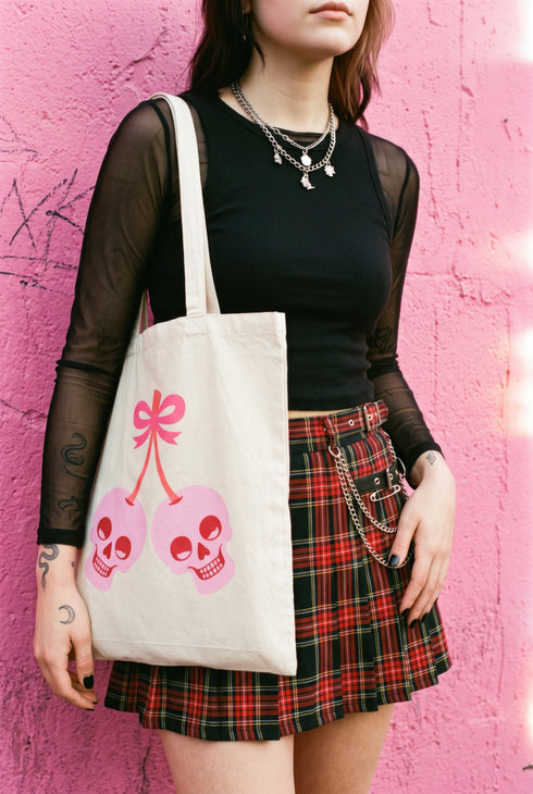 Black 15 x 16 inch 100% cotton tote bag with a kawaii pastel goth cherry-skull design printed on the front. The artwork features two soft pink skulls shaped like cherries, connected by red stems and topped with a bright pink bow. Cute, spooky, coquette, and alt-inspired everyday tote.
