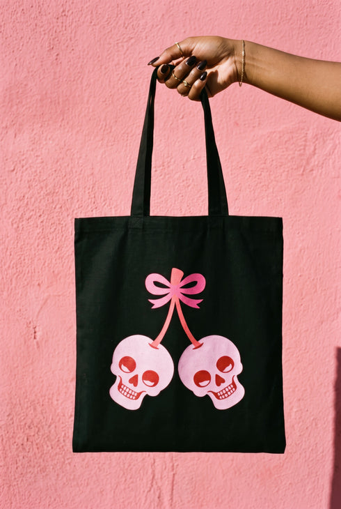 Black 15 x 16 inch 100% cotton tote bag with a kawaii pastel goth cherry-skull design printed on the front. The artwork features two soft pink skulls shaped like cherries, connected by red stems and topped with a bright pink bow. Cute, spooky, coquette, and alt-inspired everyday tote.