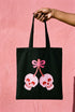 Black 15 x 16 inch 100% cotton tote bag with a kawaii pastel goth cherry-skull design printed on the front. The artwork features two soft pink skulls shaped like cherries, connected by red stems and topped with a bright pink bow. Cute, spooky, coquette, and alt-inspired everyday tote.